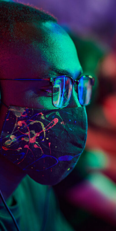 Focused young black gamer in cool mask with paint strokes pattern and eyeglasses playing online game